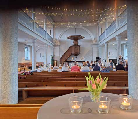 Blick in eine Kirche mit einer Abendmahlsfeier, im Vordergrund ein Tisch mit brennenden Teelichtern und einem Strauß Tulpen, im Hintergrund Menschen auf Kirchenbänken unter einer beleuchteten Decke und einer Kanzel.