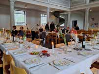 Großer festlich gedeckter Saal mit mehreren langen Tafeln, an denen Menschen sitzen und stehen, sichtbar bei einer Abendmahlsfeier am Gründonnerstag.