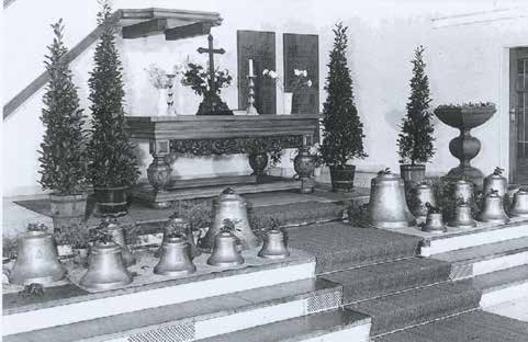 Schwarz-weiße Aufnahme eines Altars mit mehreren Glocken unterschiedlicher Größe auf Stufen davor, flankiert von hohen Pflanzen in Töpfen, im Hintergrund ein Kreuz und Kerzen auf dem Altar