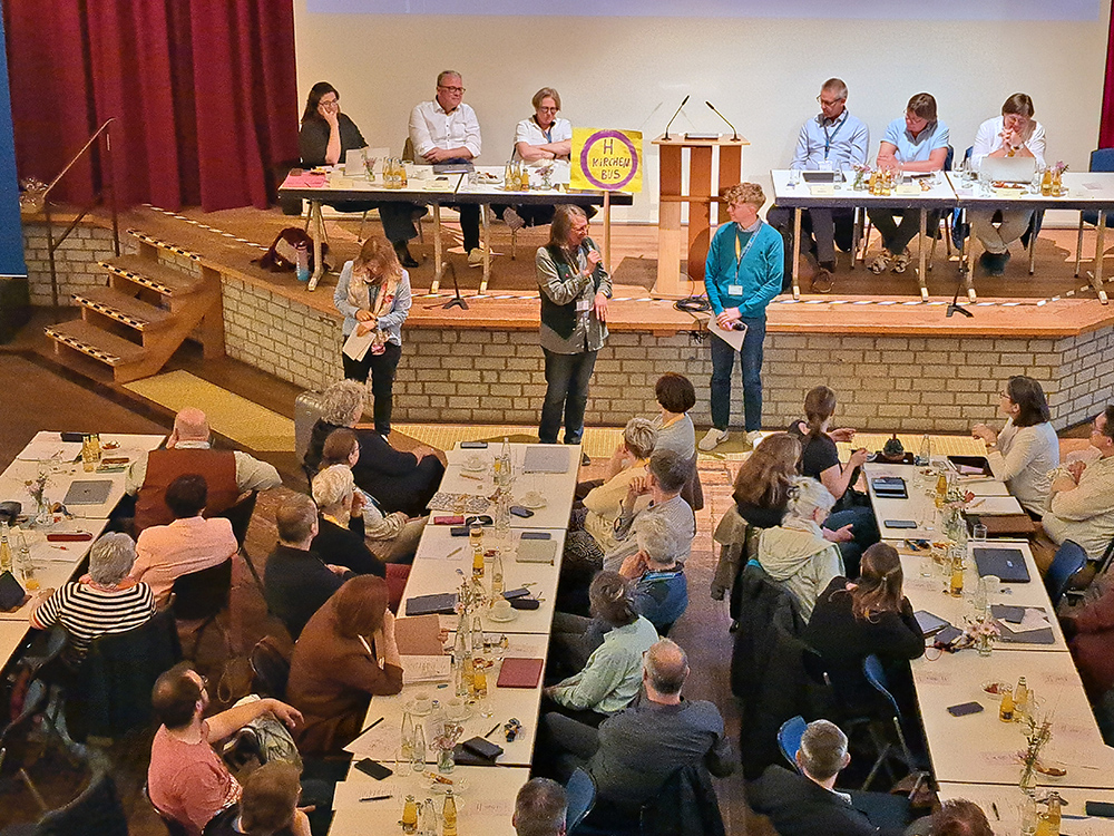 Versammlung der Synode des Kirchenkreises mit mehreren Personen an Tischen und einem Podium im Hintergrund, auf dem sechs Personen sitzen, eine Person spricht mit Mikrofon in der Mitte des Raums