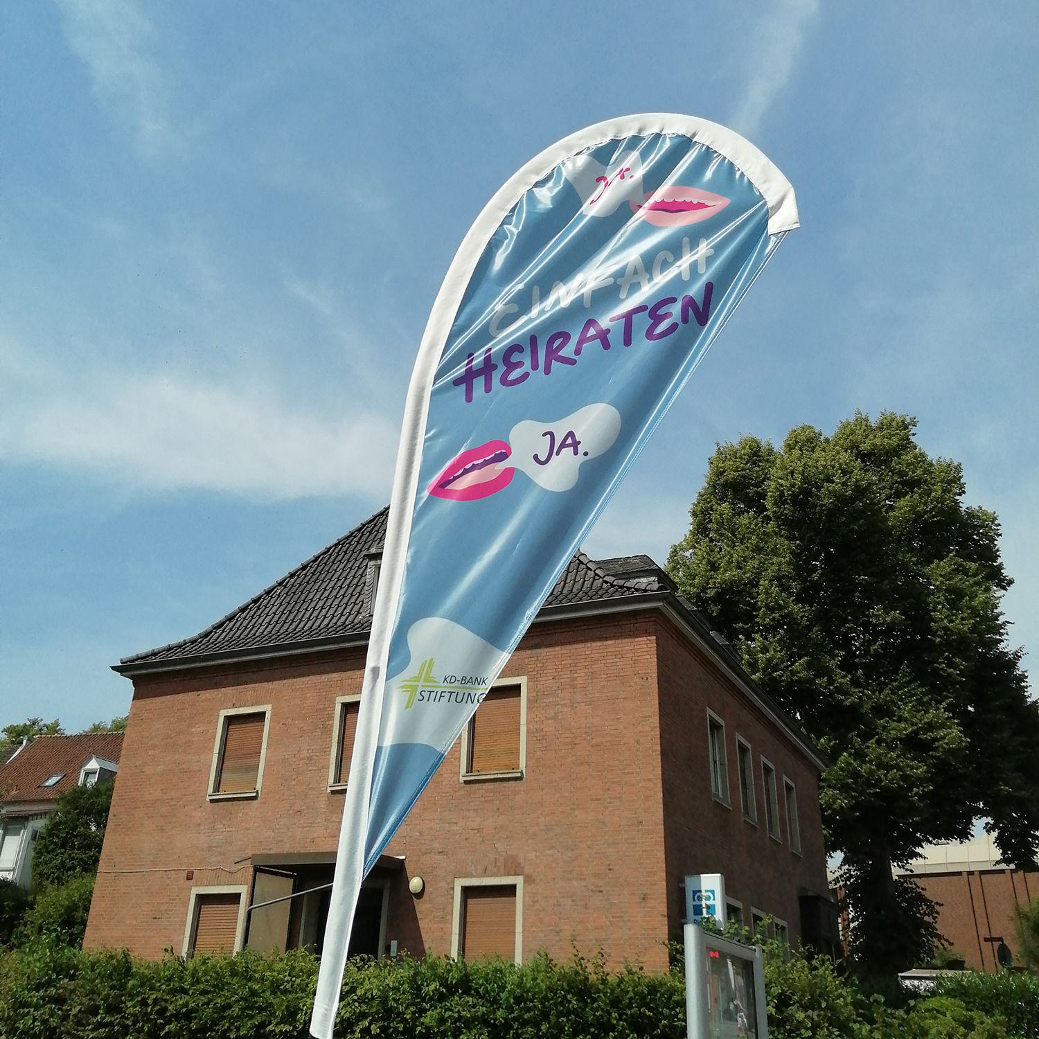 Blaues Wimpelbanner mit der Aufschrift 'Einfach heiraten' und zwei roten Lippenstiftabdrücken vor einem Backsteinhaus und blauem Himmel