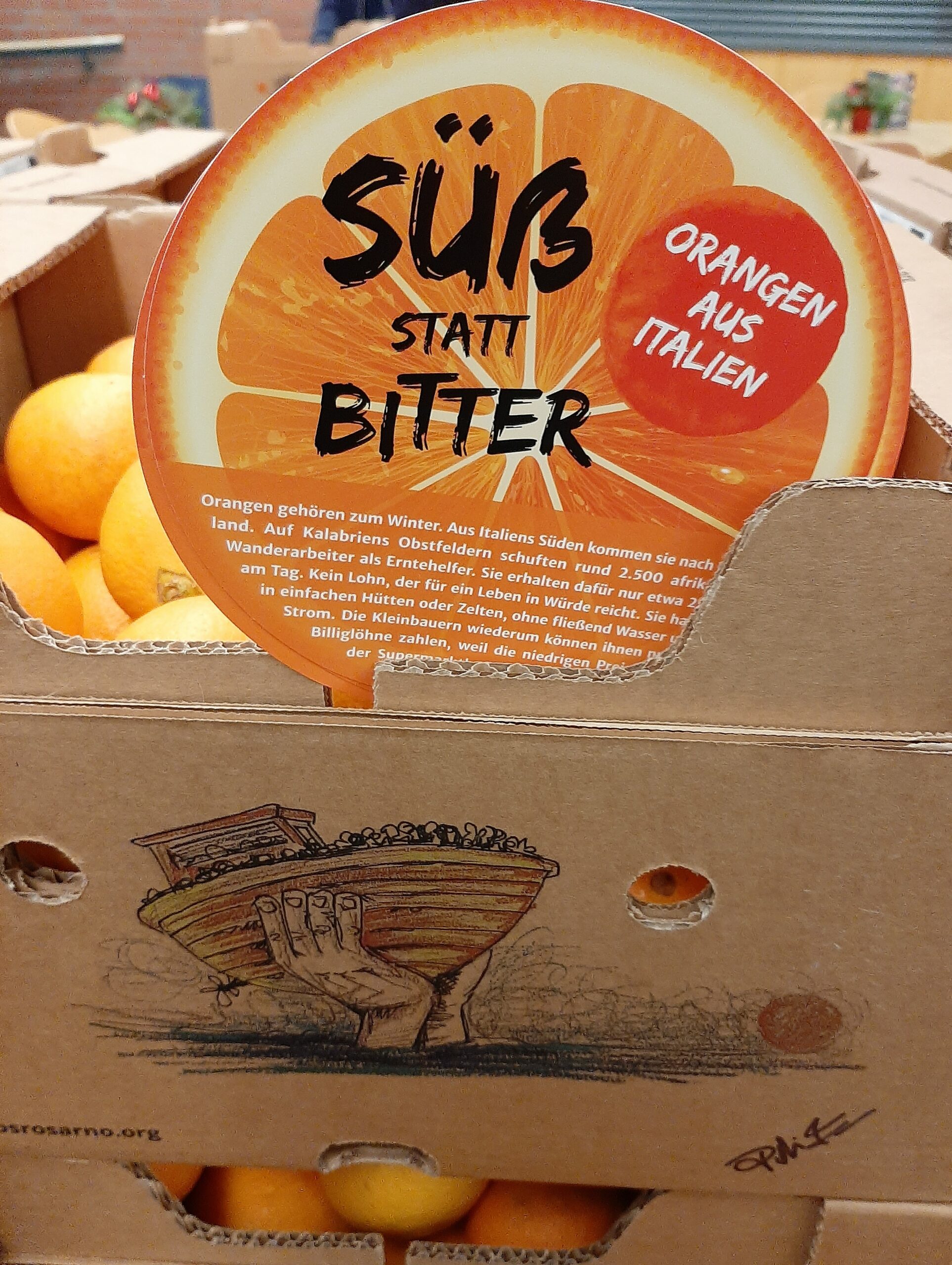 Orangenschachtel mit Aufkleber 'SÜß STATT BITTER ORANGEN AUS ITALIEN' und Text über Orangenanbau sowie Illustration einer Hand, die ein Boot hält